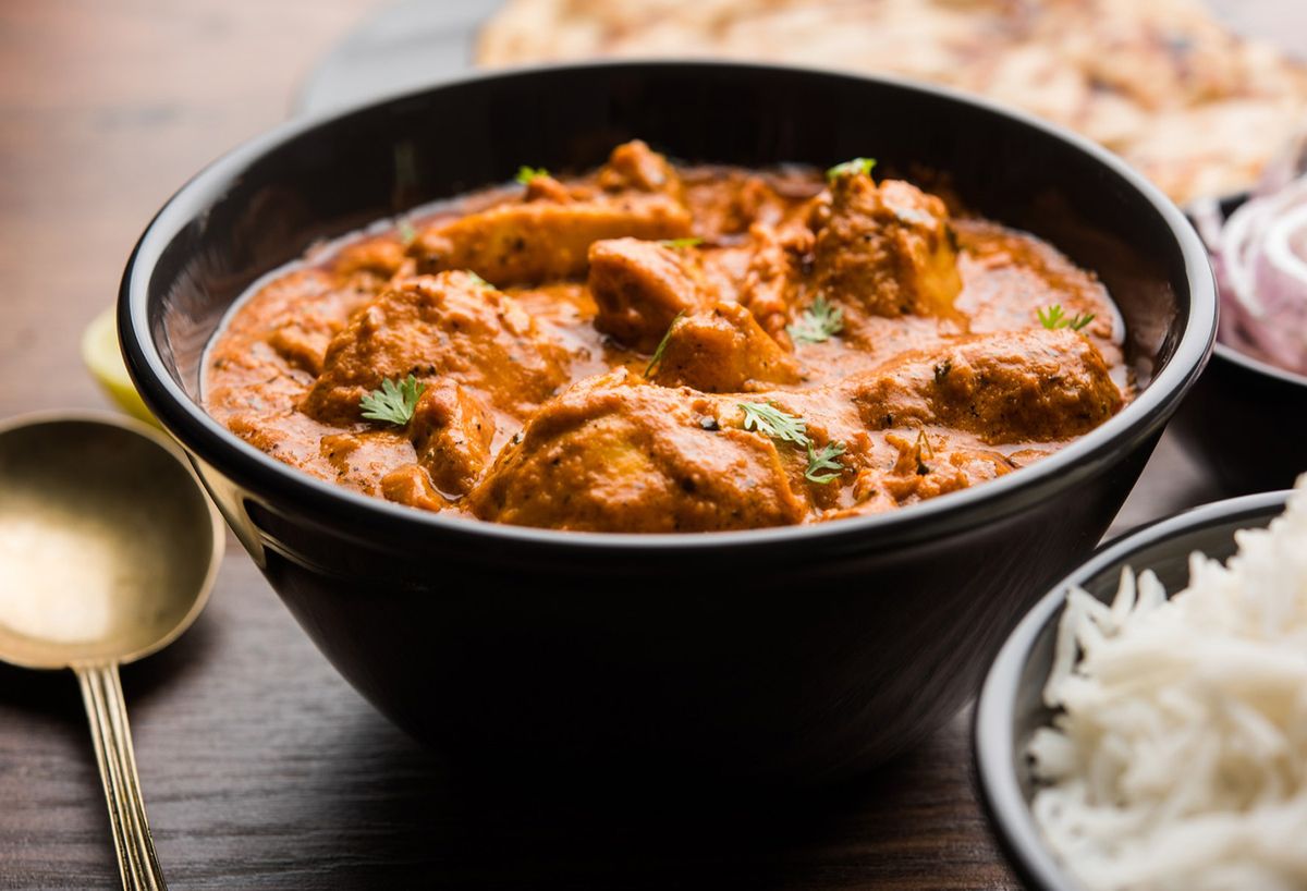 Close-up of a bowl of creamy chicken curry garnished with fresh cilantro, served alongside steamed rice, naan, and sliced onions on a wooden table.