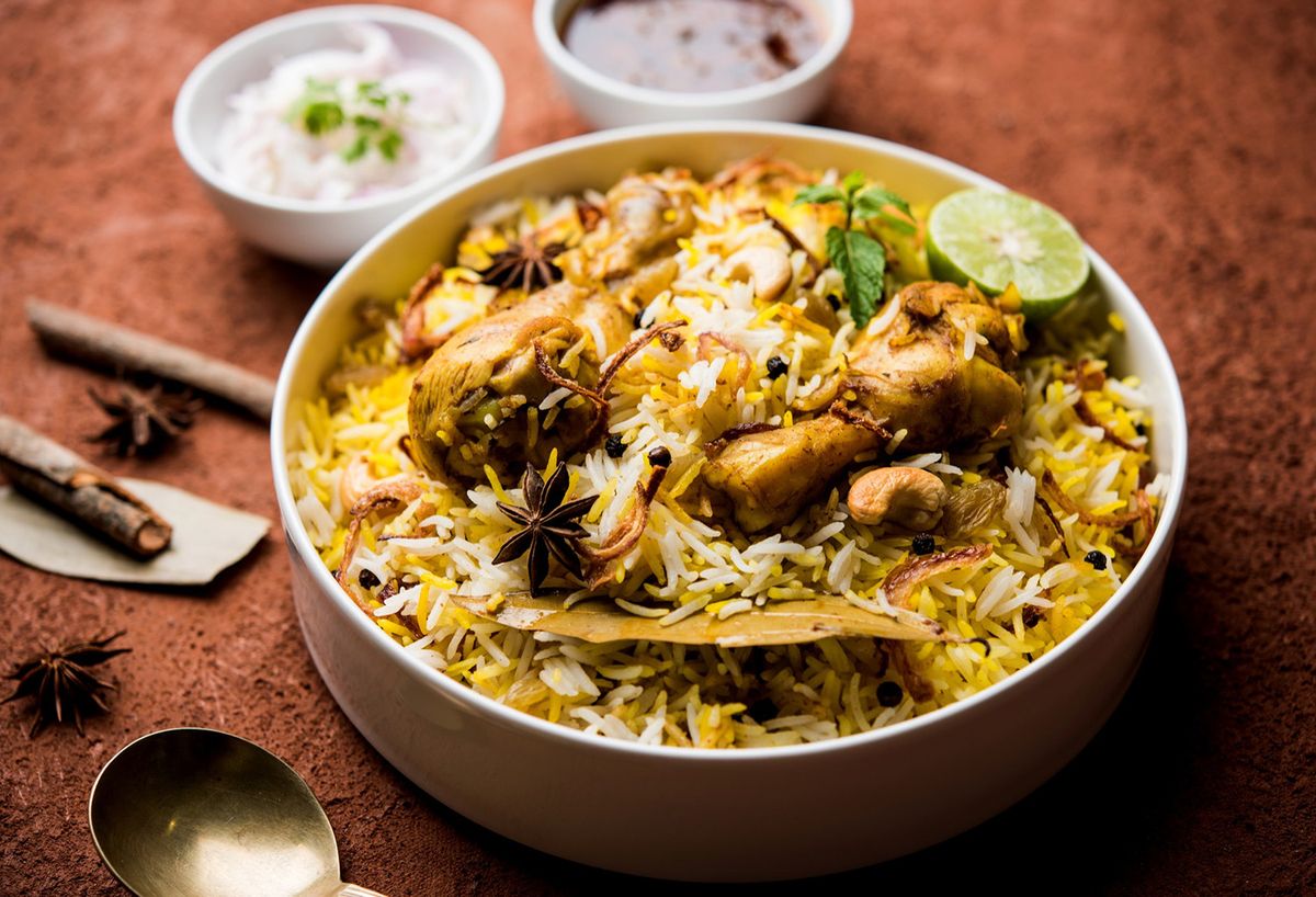 Bowl of aromatic chicken biryani made with long-grain basmati rice, tender chicken, fried onions, whole spices, and cashews, served with raita, curry, and a fresh lime wedge.