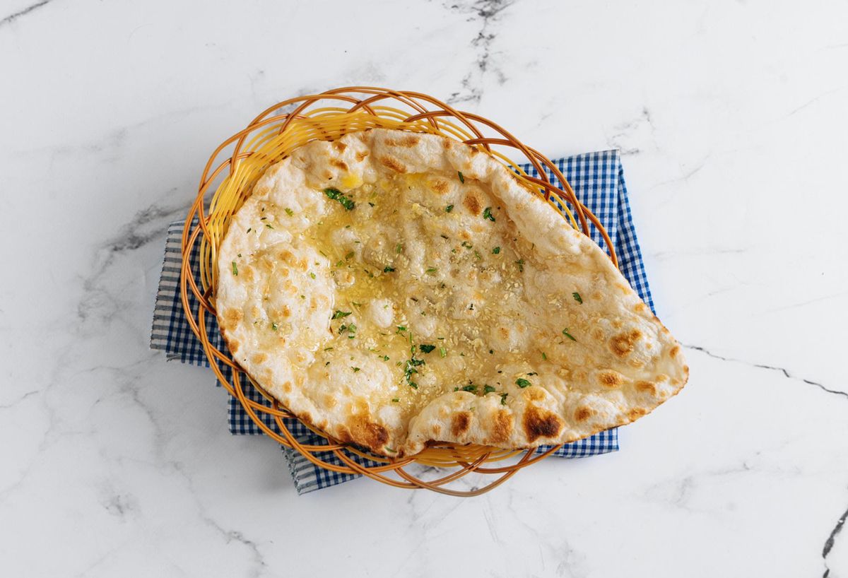 Fresh garlic naan brushed with butter and topped with minced garlic and herbs, served in a woven basket on a marble tabletop.