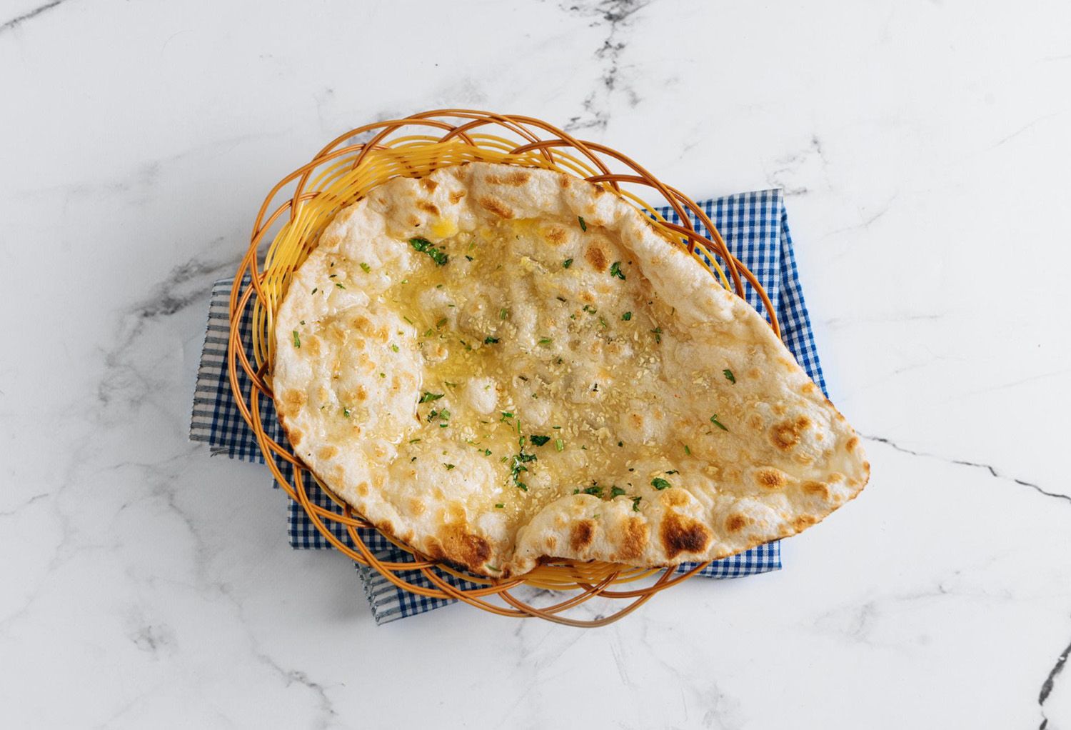 Fresh garlic naan brushed with butter and topped with minced garlic and herbs, served in a woven basket on a marble tabletop.