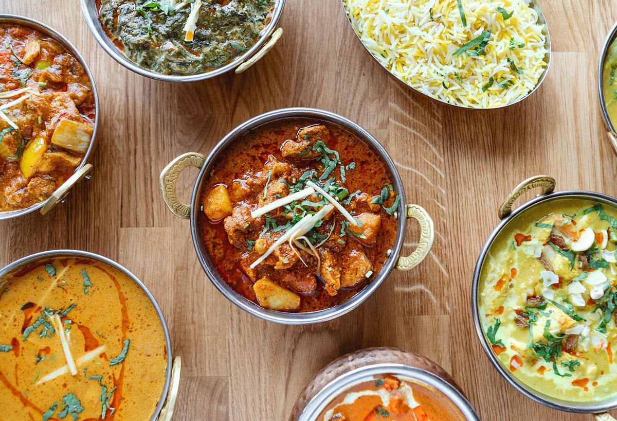 Overhead view of assorted curry dishes served in traditional bowls with basmati rice on a wooden table, featuring a variety of rich gravies garnished with fresh herbs and spices.